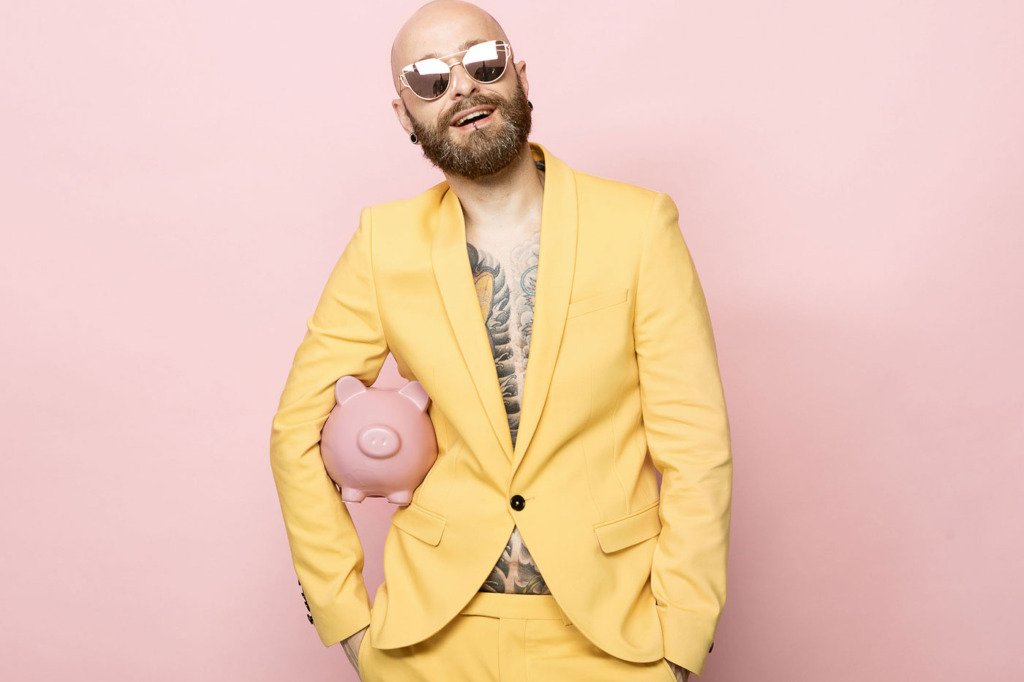 Mann im gelben Anzug steht vor einem rosanen Fotohintergrund und hält ein Sparschwein